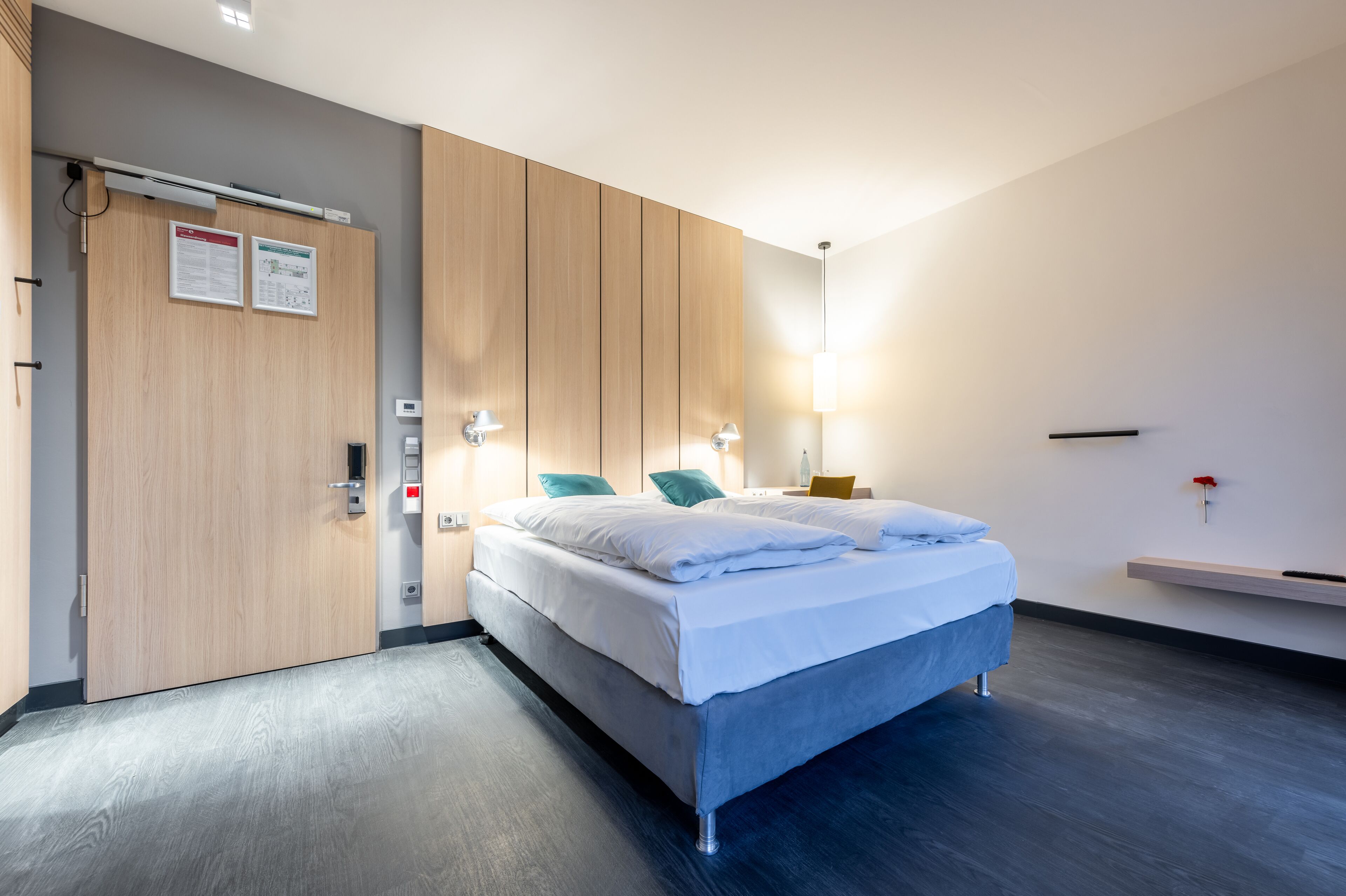 Ein modernes Hotelzimmer mit Bett, Holzschrank, Nachttisch und einer kleinen Ablagefläche.