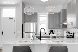 A modern kitchen with grey cabinets, a stainless steel refrigerator, a black faucet, and pendant lights.