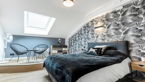 A bedroom with a skylight, a bed with a dark cover, a nightstand, a chair, and a patterned wall.