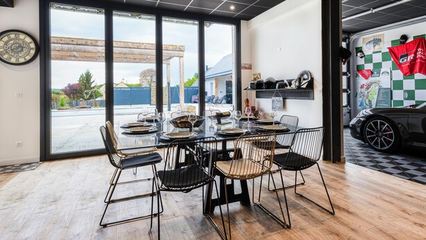 A modern dining area with a glass table and metal chairs, a wooden ceiling, and a view of a garage with a car and racing memorabilia.