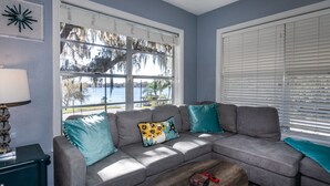 A living room with a gray sectional sofa, teal and yellow patterned pillows, a wooden coffee table, and a view of a lake through large windows.