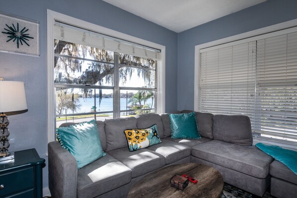 A living room with a gray sectional sofa, teal and yellow patterned pillows, a wooden coffee table, and a view of a lake through large windows.