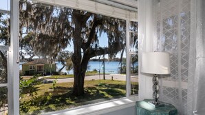 A room with a large window overlooking a lake, a lamp, and a view of a house and trees.