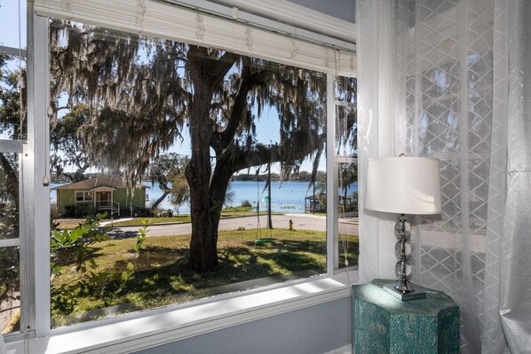 A room with a large window overlooking a lake, a lamp, and a view of a house and trees.