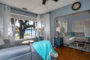 A living room with a sofa, a coffee table, and a side table. There is a window with curtains and a view of a tree and water outside.