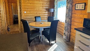 L’intérieur d’un chalet en bois comprend une table à manger ronde, quatre chaises, un coin cuisine et une fenêtre avec des rideaux.