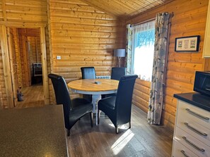 A wooden cabin interior with a round dining table, four chairs, a kitchen area, and a window with curtains. - Chestnut Meadow Cabin (East Sussex)