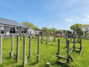 A playground with wooden structures and a fenced area, a building with windows, and a clear sky.