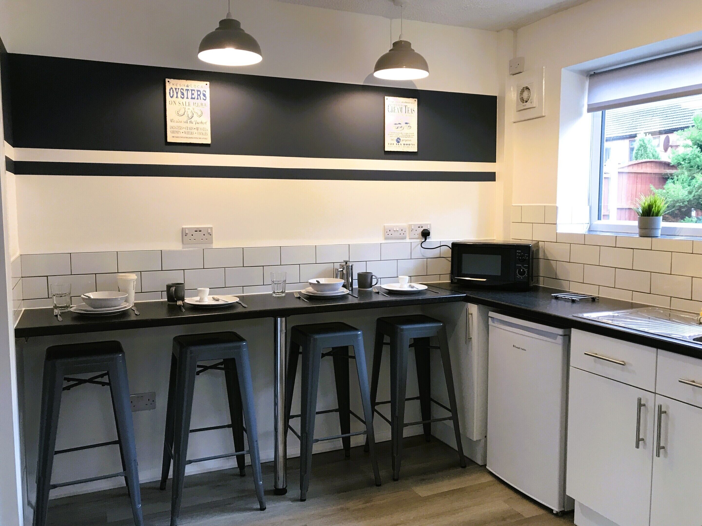 A kitchen with a black countertop, white cabinets, and two bar stools. There are signs on the wall advertising oysters and cream teas.
