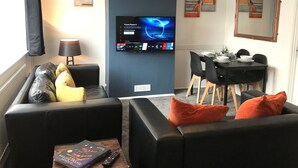 A modern living room with a flat-screen TV mounted on a dark wall, a black leather sofa with orange cushions, a dining table with chairs, and a lamp on a side table. - Modern 3-bed House With Parking Bamber Bridge, Preston (Preston)