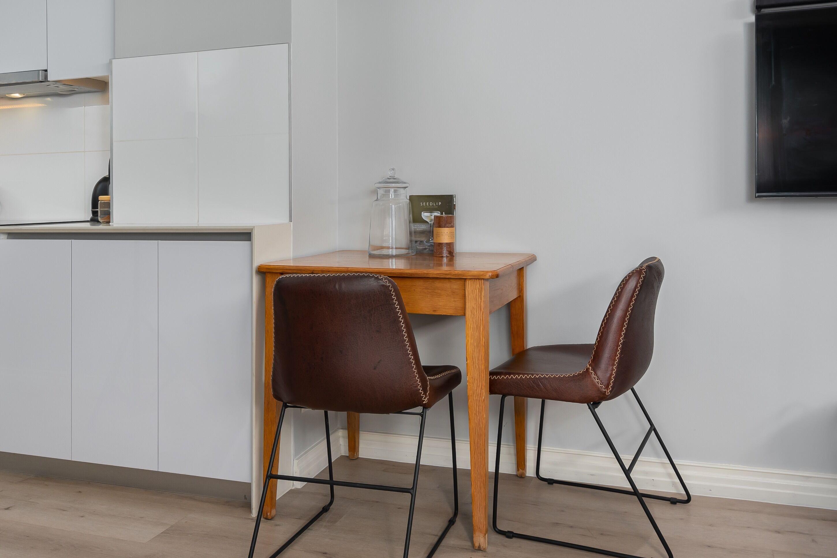 Une cuisine moderne avec des meubles blancs, une table en bois, deux chaises en cuir marron et un récipient en verre posé sur la table.