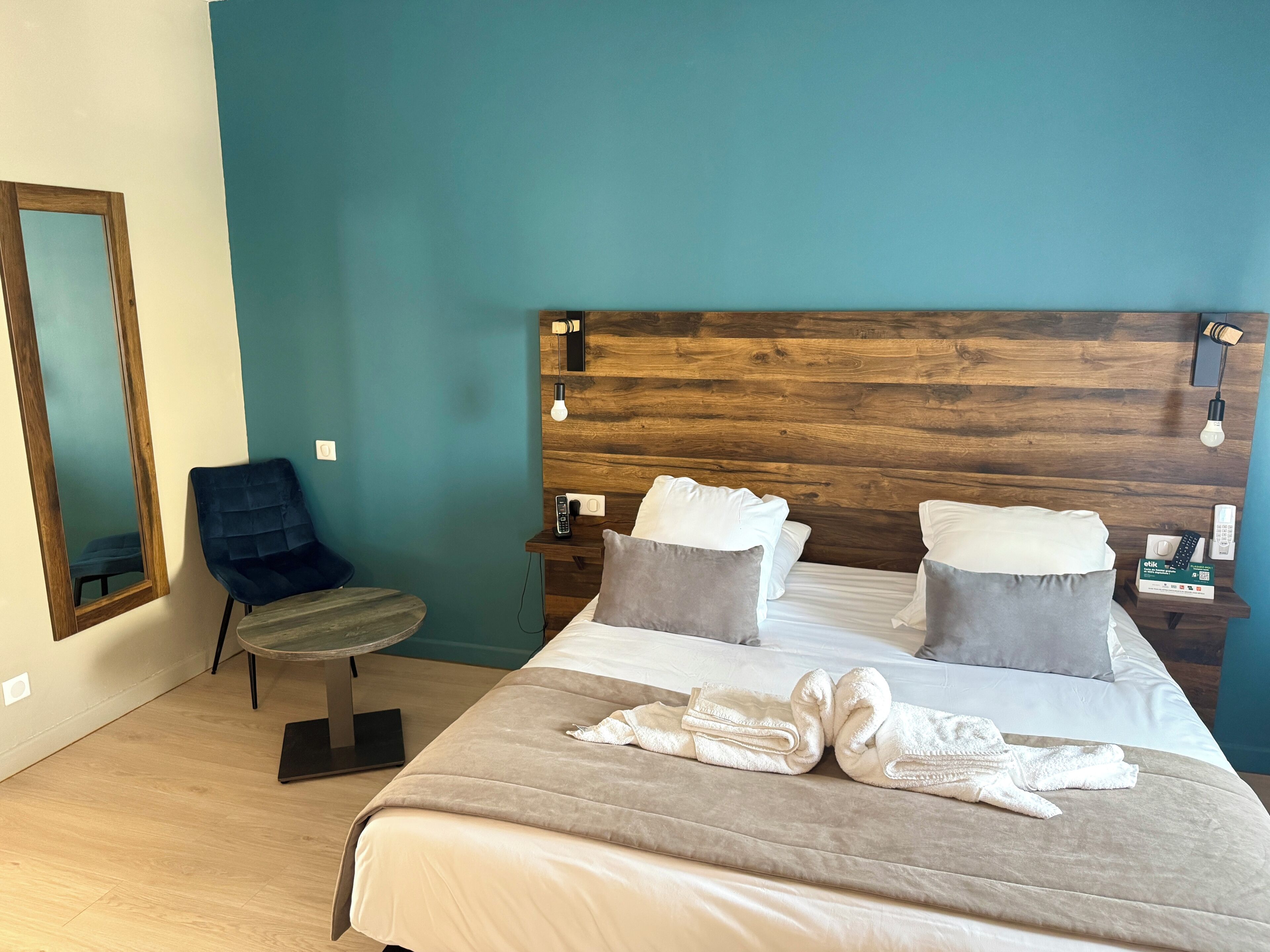 Une chambre d’hôtel avec une tête de lit en bois, un lit avec du linge de lit blanc, deux oreillers, une table de chevet avec une lampe, une chaise et une table ronde.