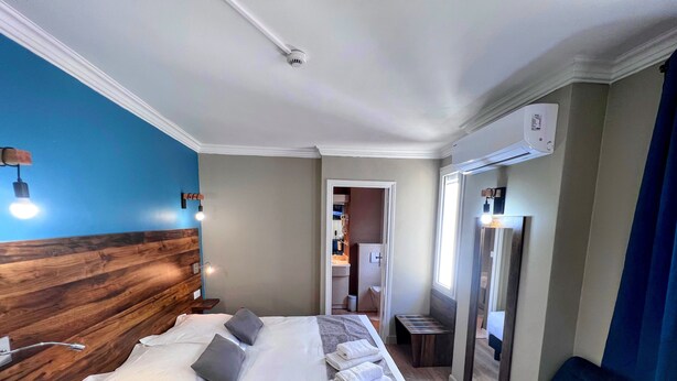 A hotel room with a wooden headboard, a large bed with white linens, a blue chair, and a bathroom visible through an open door. - Logis Hôtel de la Poste (Vitry-le-Francois)