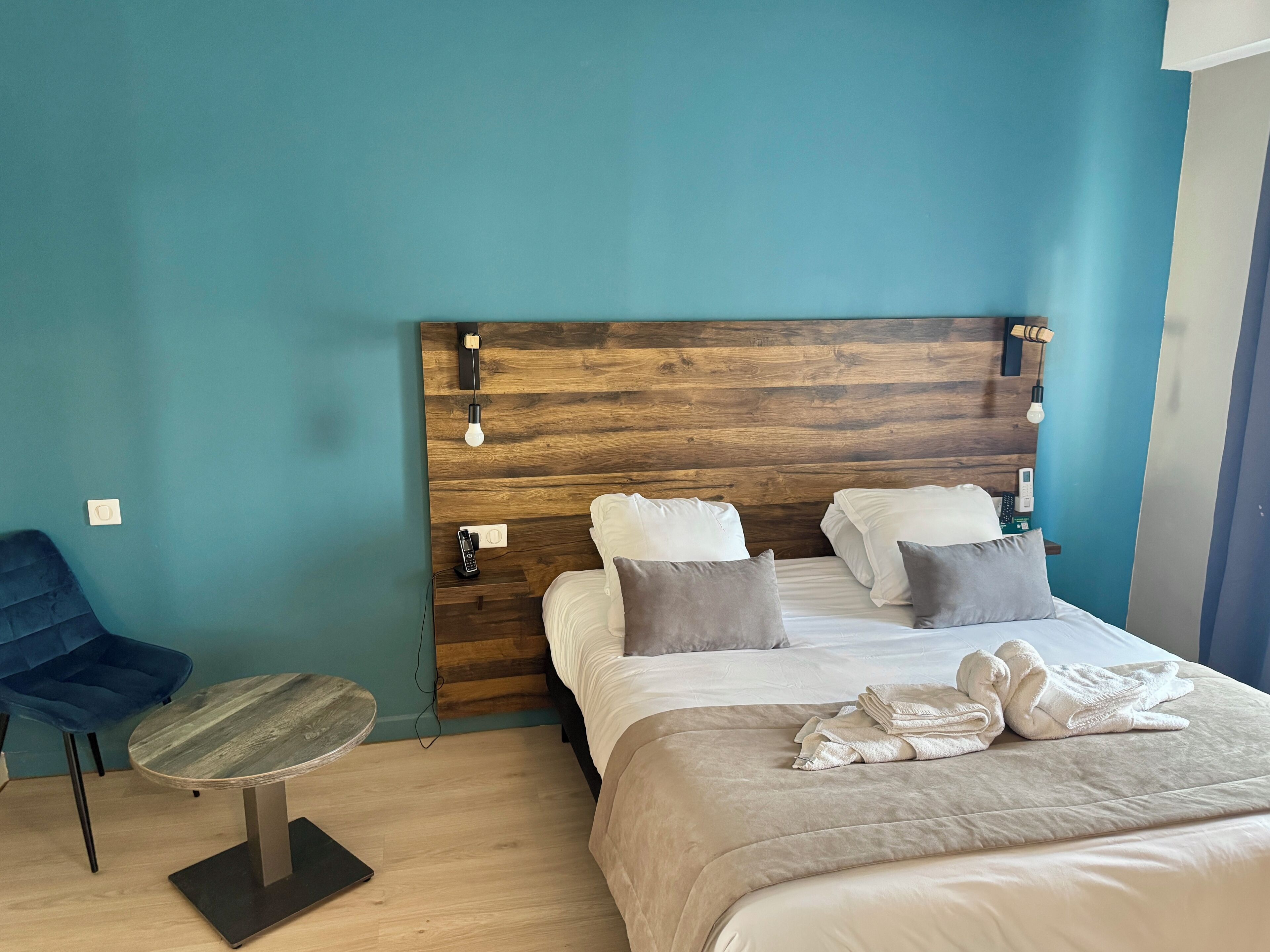 Een hotelkamer met een houten hoofdeinde, een bed met kussens, een blauwe stoel en een kleine ronde tafel.