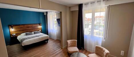 A hotel room with a bed, a round table, two chairs, a TV, and a window with curtains.