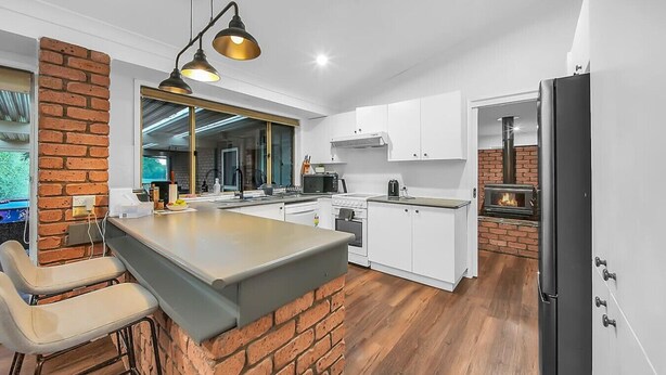 A modern kitchen with a brick accent wall, a central island, white cabinetry, and a built-in wood stove. - Private House - Pool, Games and Fire Pit Bathurst (Robin Hill)