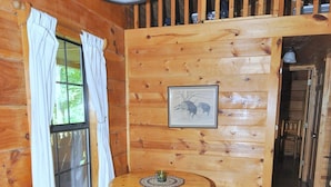 A wooden interior with a round table and benches, a framed picture on the wall, and a window with curtains. - Sleepy Creek - Creekside Escape (Berkeley Springs)