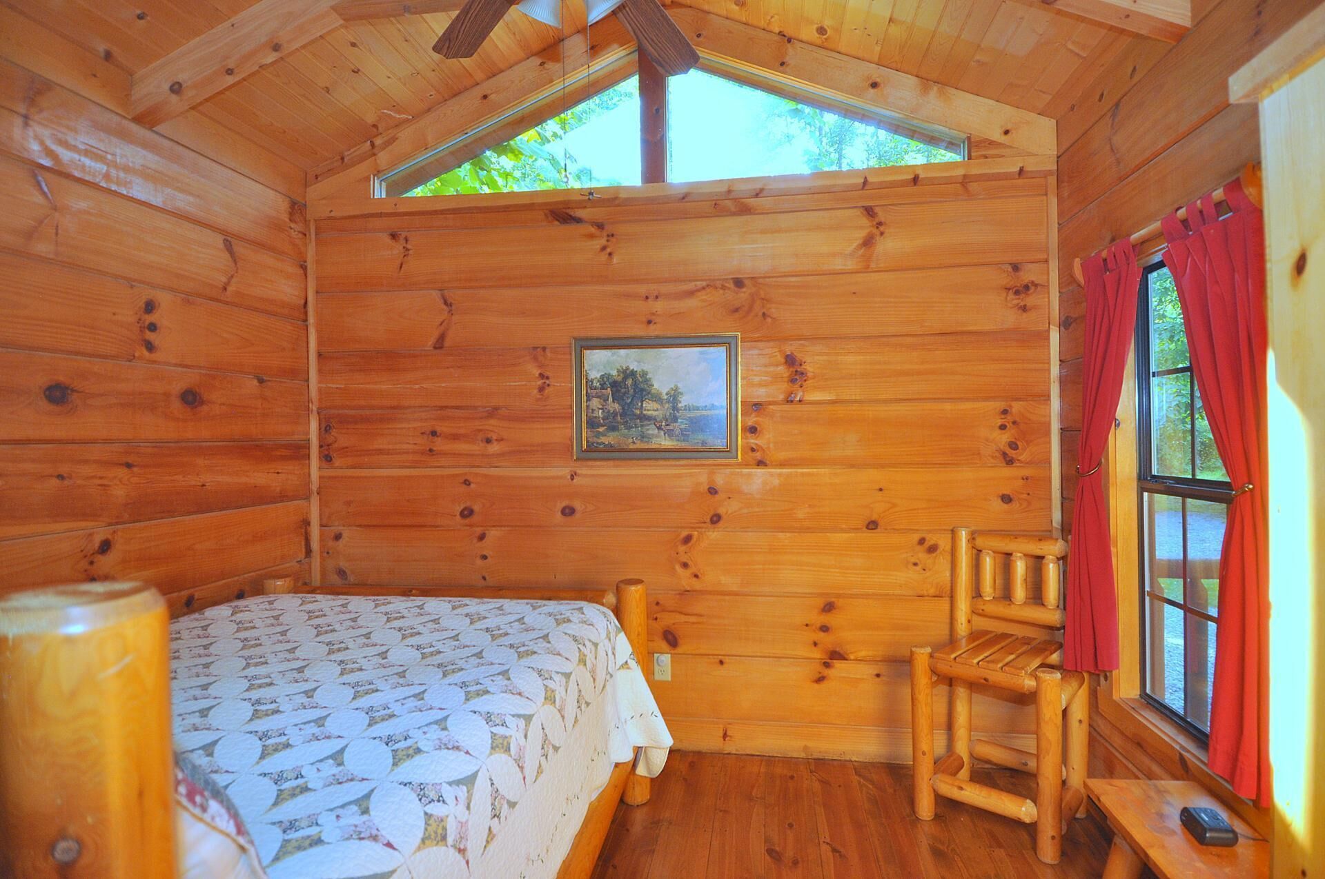A wooden cabin room with a bed, a chair, a table, a painting on the wall, and a window with curtains.