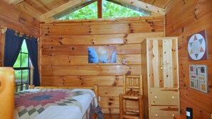 A cozy cabin room with a bed, a wooden chair, a cabinet, and a window with a view of greenery. - Sleepy Creek - Creekside Escape (Berkeley Springs)