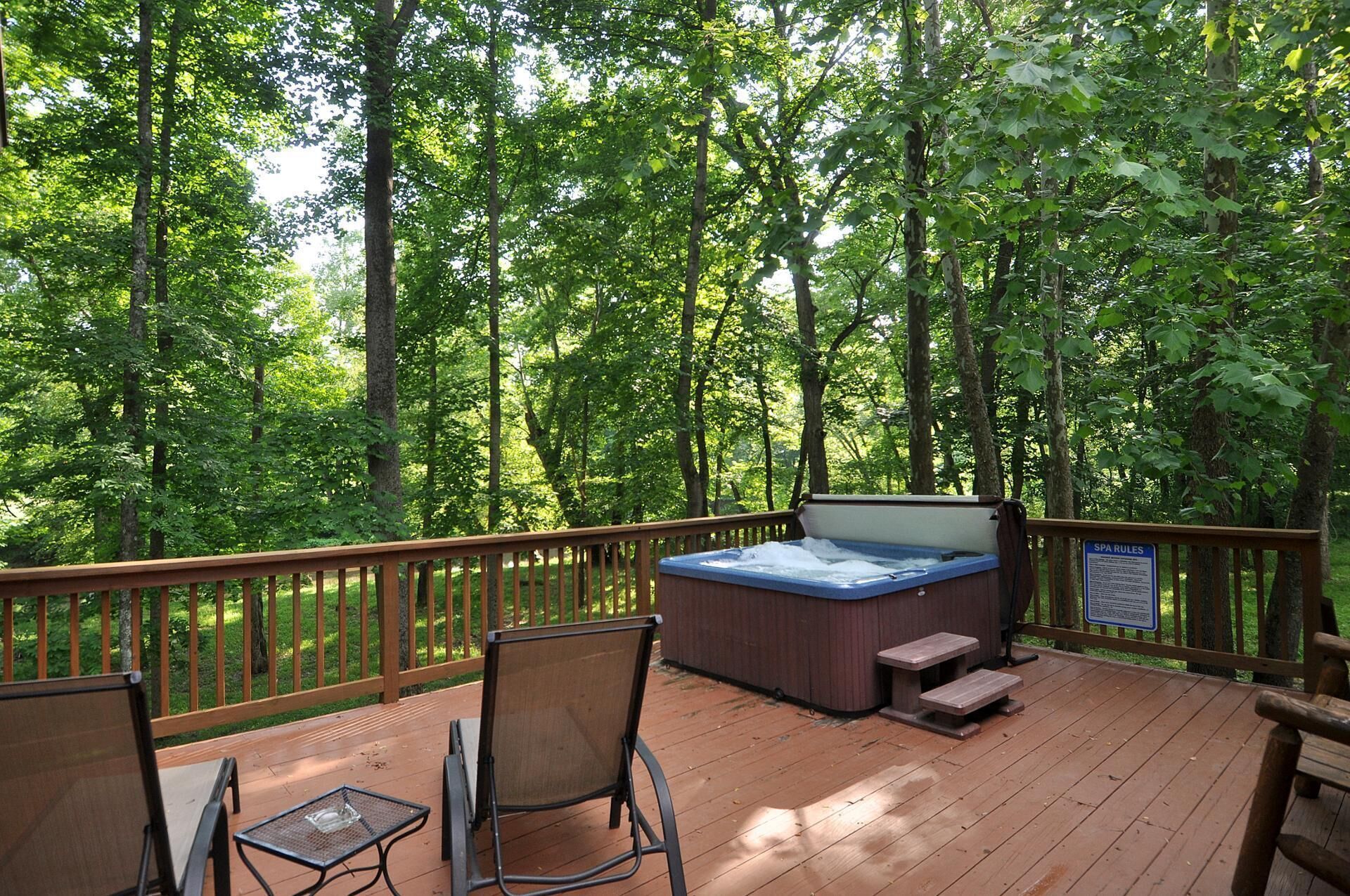 A wooden deck with a hot tub, two chairs, and a table, surrounded by a forest.