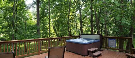 A wooden deck with a hot tub, two chairs, and a table, surrounded by a forest.