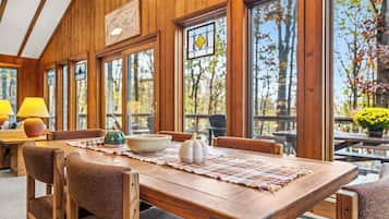 A wooden interior with a dining table, chairs, and large windows offering a view of trees and outdoor seating.