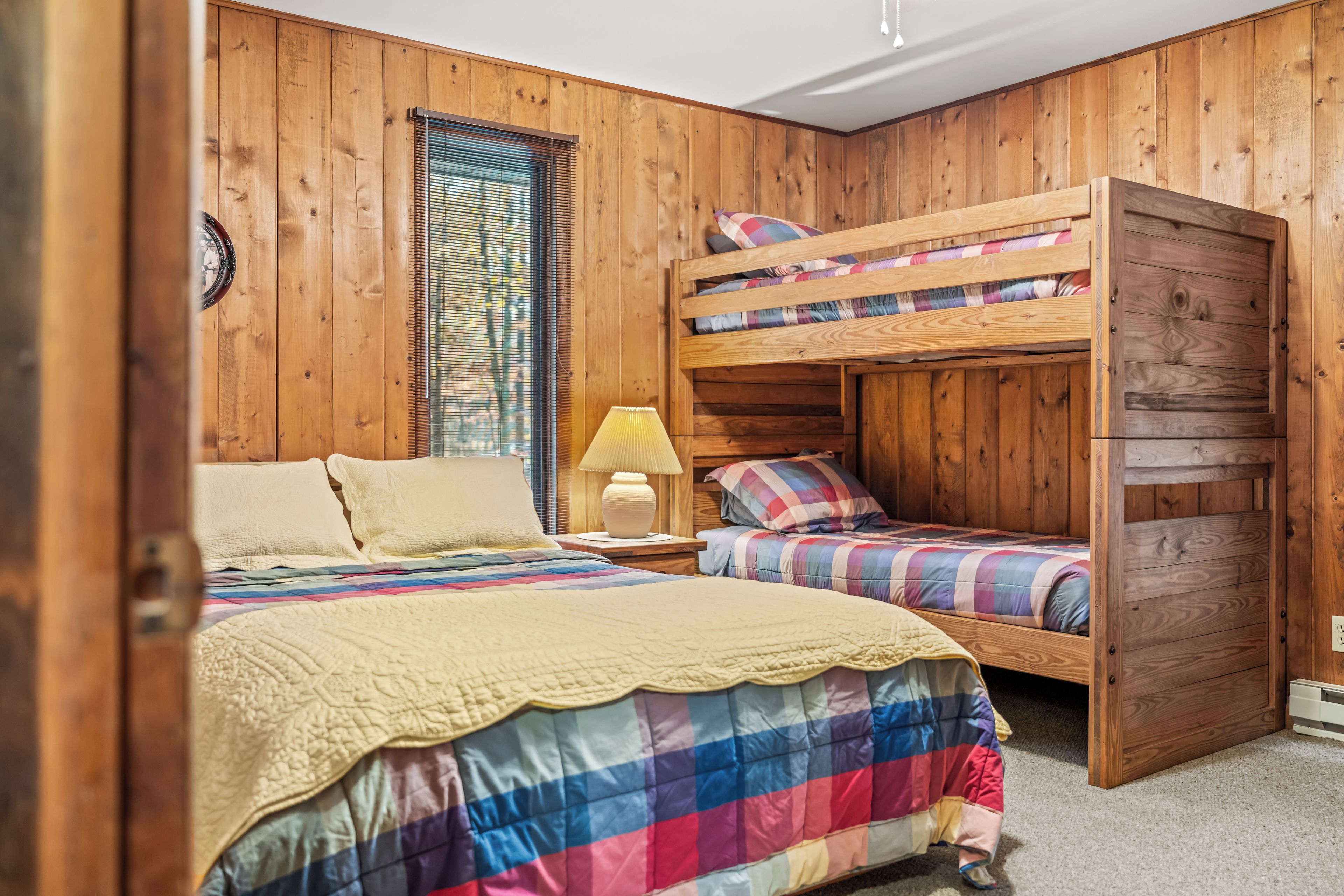 A room with wooden walls, a bunk bed, a single bed, and a window with blinds.