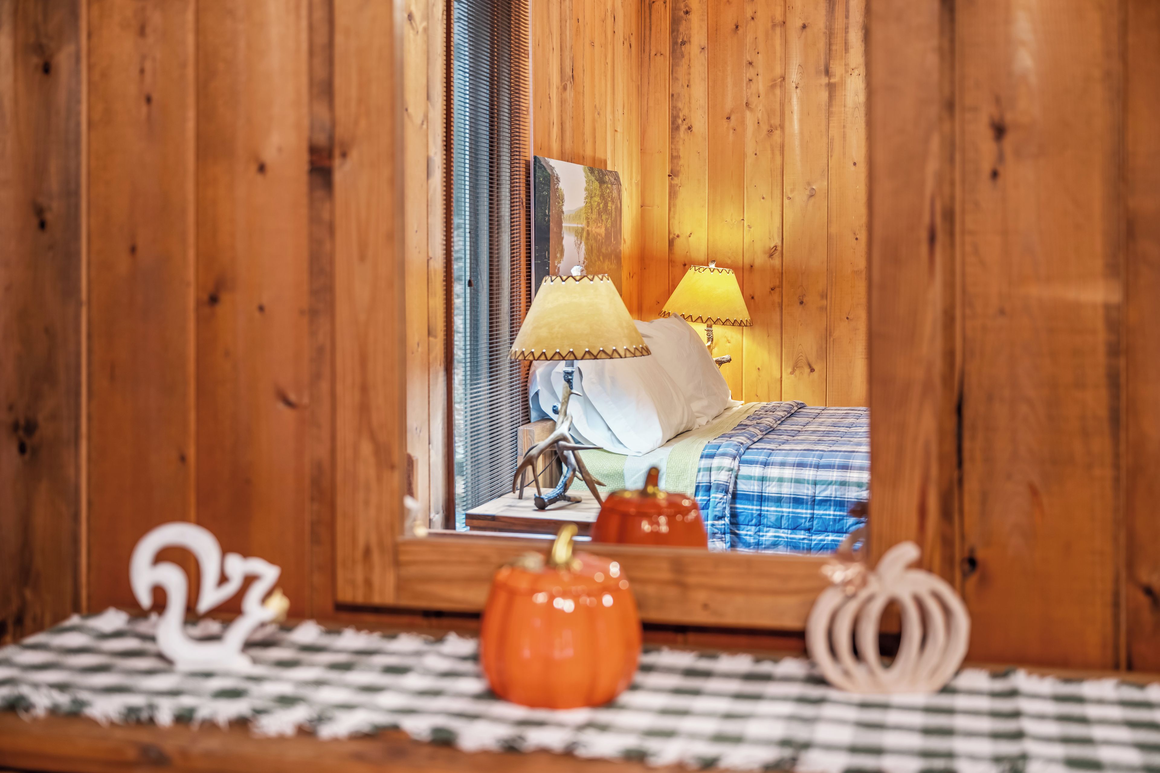 Interior de madera con una cama, dos lámparas, una mesita con objetos decorativos y un espejo que refleja la habitación.