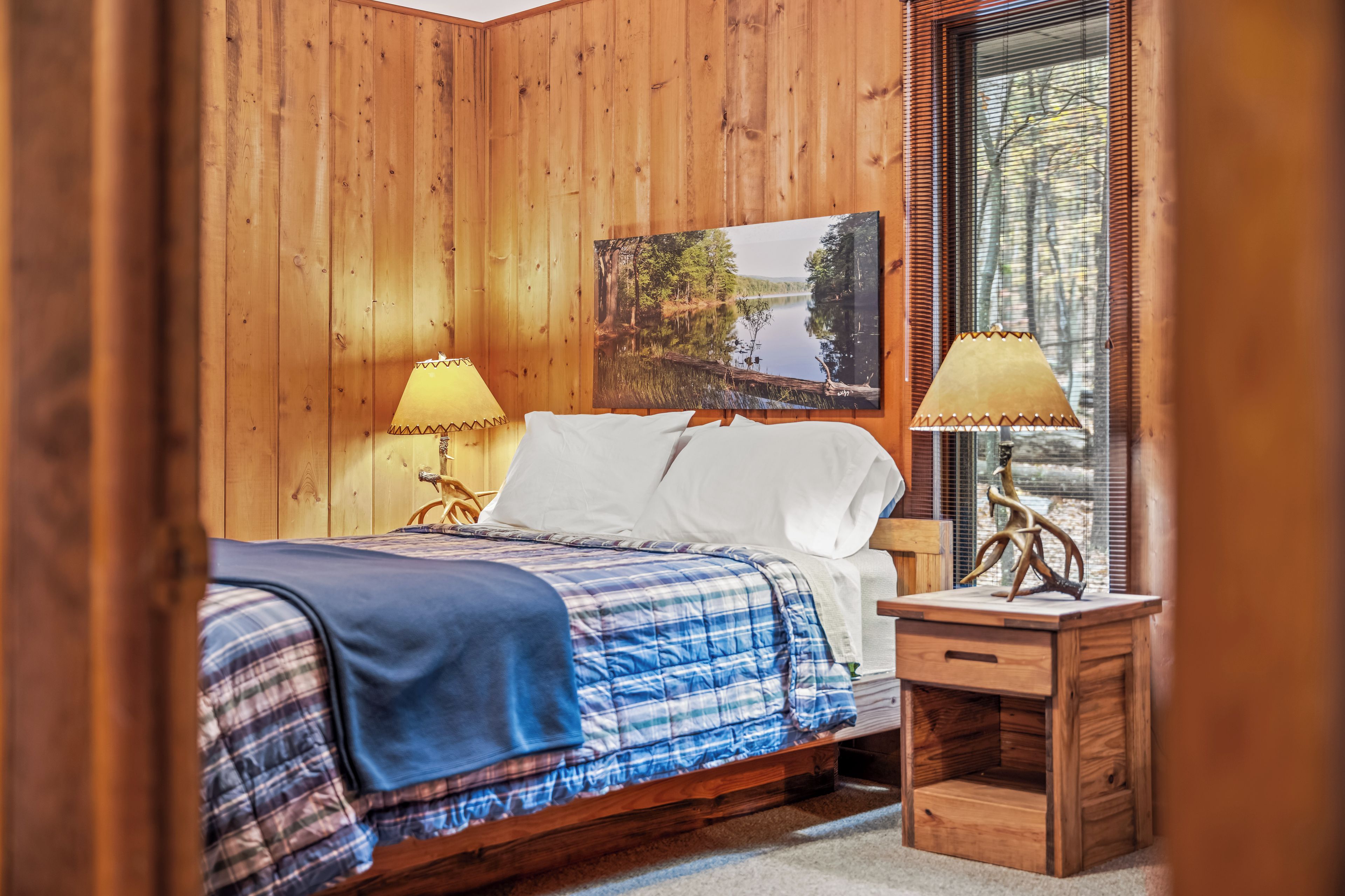 A wooden-paneled room with a bed, two bedside lamps, a nightstand, and a window with blinds.
