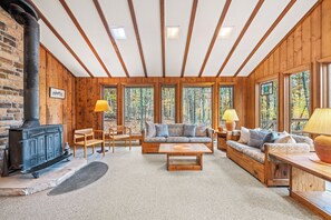 Interior acogedor de una cabaña con chimenea, muebles de madera y grandes ventanales que dan a un bosque.