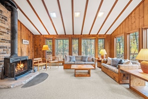 Interior acogedor de una cabaña con chimenea, muebles de madera y ventanas amplias con vista al bosque.