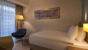 Une chambre d’hôtel avec un lit, un bureau, une chaise, une lampe et une fenêtre avec des rideaux.