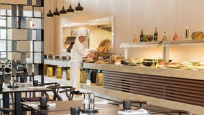 A modern restaurant interior with a chef preparing food at the counter, tables with chairs, and a variety of condiments and dishes displayed on shelves. - The Superior Living Batisehir  (Istanbul)