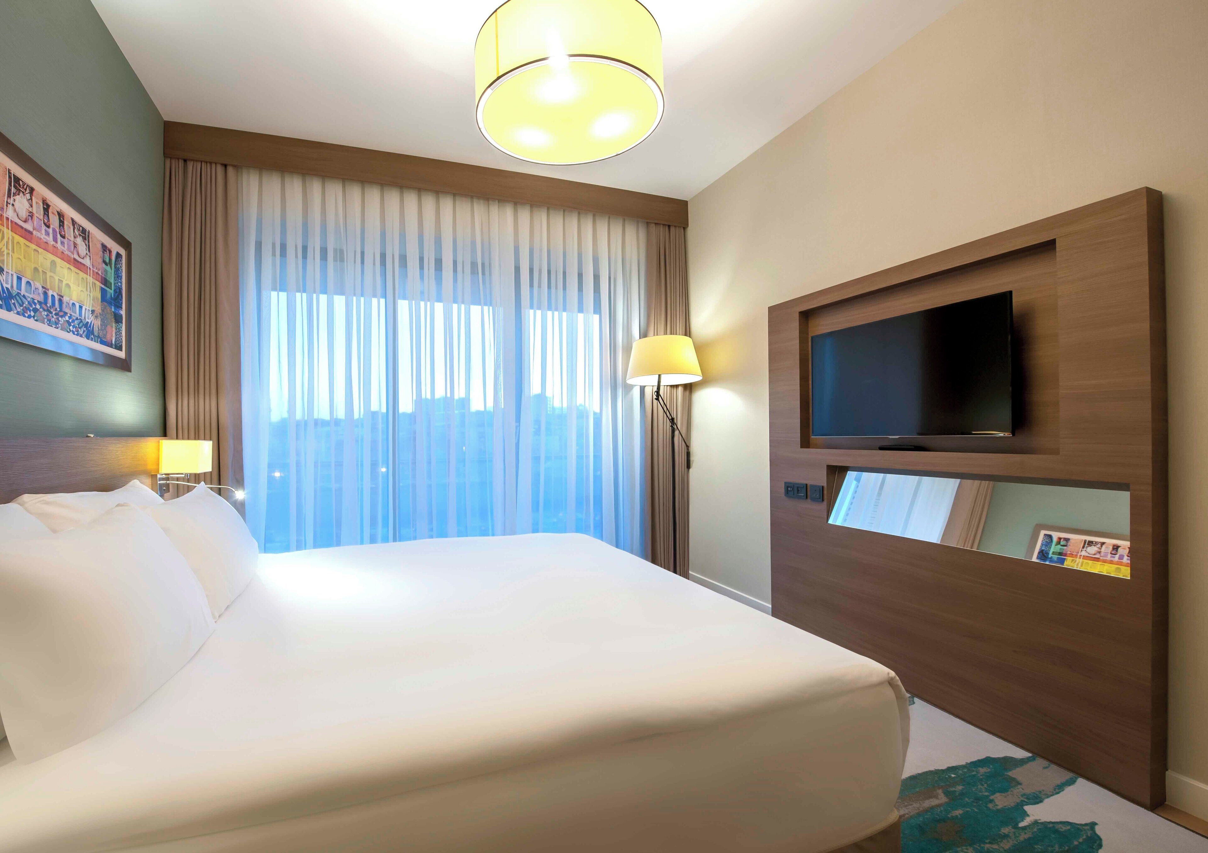 Habitación de hotel con una cama grande, un televisor de pantalla plana montado en la pared, una ventana con cortinas traslúcidas y un cuadro colgado.