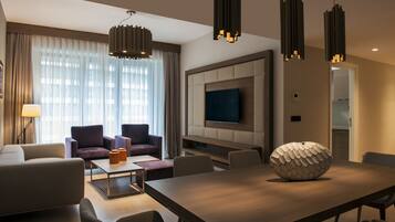Una habitación de hotel moderna con un televisor de pantalla plana grande montado en la pared, una mesa de comedor de madera con un original centro de mesa decorativo, y un área de descanso con sofás y una mesa de centro.