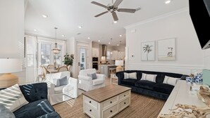 A spacious living area with a ceiling fan, a dark-colored sofa, a white armchair, a wooden coffee table, and a kitchen in the background.