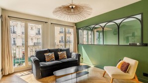 A modern living room with a black sofa, a white armchair, a wooden floor, a large window with a view of buildings, and a unique ceiling light fixture.