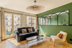 A modern living room with a black sofa, a white armchair, a wooden floor, a large window with a view of buildings, and a unique ceiling light fixture. - Stylish 3BD for 10 guests next to Invalides! (Paris)
