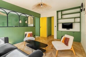 En moderne stue med sofa, lænestol og sofabord. Et fjernsyn er monteret på væggen, og en dør fører til et andet rum.