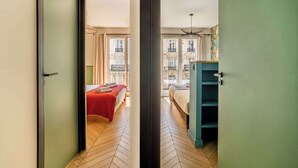 A hotel room with a herringbone floor, a bed with a red bedspread, a blue wardrobe, and a balcony with a view of buildings.