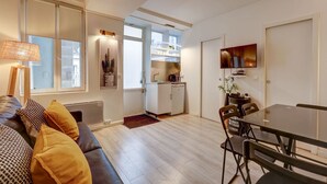 A modern living room with a sofa, a dining table with chairs, a kitchen area with a refrigerator, and a TV mounted on the wall. - 2 bedroom in the heart of Le Marais! (Paris)