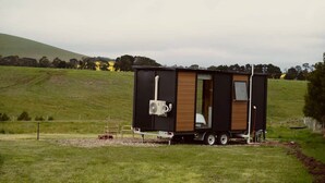 A small, modern mobile home with a wooden interior, situated on a grassy field with a hilly background. - Maluridae Creek Views by Tiny Away (Lawrence)