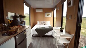 A compact, modern room with a bed, bedside table, and a small desk with a chair. There is a window providing a view of the outdoors. - Maluridae Creek Views by Tiny Away (Lawrence)