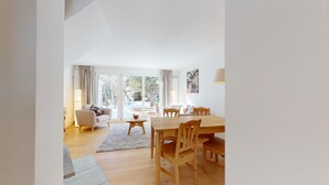 A spacious living area with a wooden dining table, chairs, a sofa, and a coffee table. There is a sliding glass door leading to an outdoor area with a view of trees and a garden.