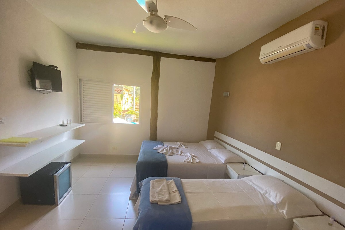 A hotel room with two single beds, a ceiling fan, an air conditioner, a window with blinds, a small TV on a shelf, and a trash bin.