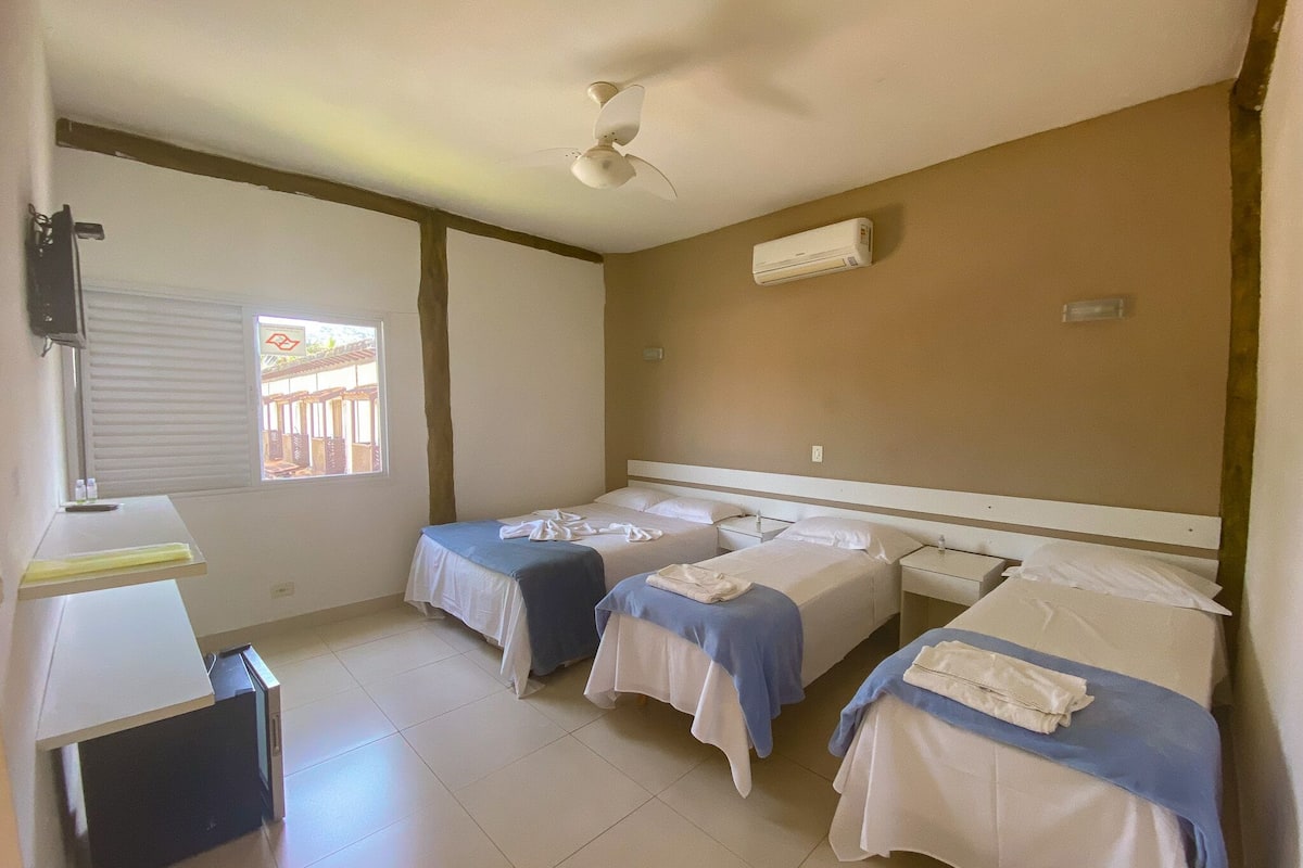A hotel room with three single beds, a ceiling fan, an air conditioning unit, and a window with blinds.