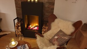 A cozy room with a fireplace, a leather armchair with a fluffy pillow, a glass candle holder, and a magazine on a wooden table. - 3-room apartment on the first floor, 67m2. (Celerina/Schlarigna)
