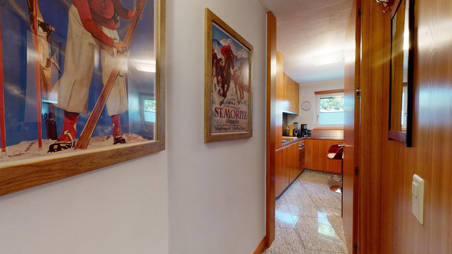 A hallway leading to a kitchen with wooden cabinets, a bar stool, and a poster of a horse race.