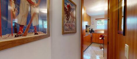 A hallway leading to a kitchen with wooden cabinets, a bar stool, and a poster of a horse race.