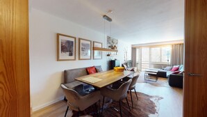 A modern living room with a wooden dining table, leather chairs, and a sofa. There are framed pictures on the wall, a large window with curtains, and a door to the right. - 3-room apartment on the first floor, 67m2. (Celerina/Schlarigna)